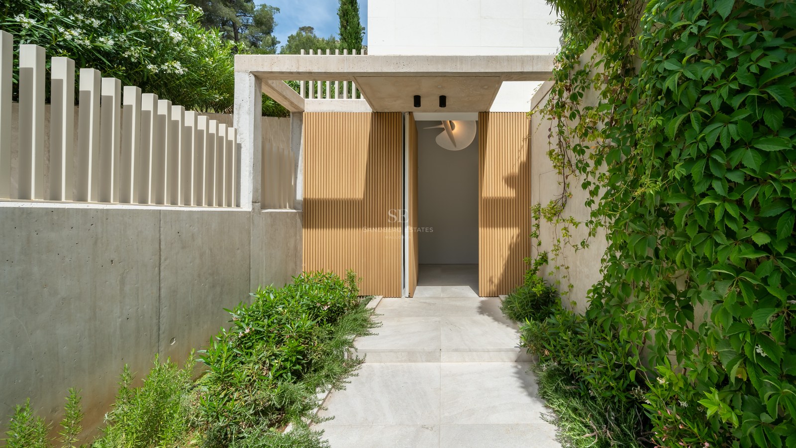 Entrada de villa moderna con listones de madera, muros de hormigón visto y exuberante vegetación mediterránea.