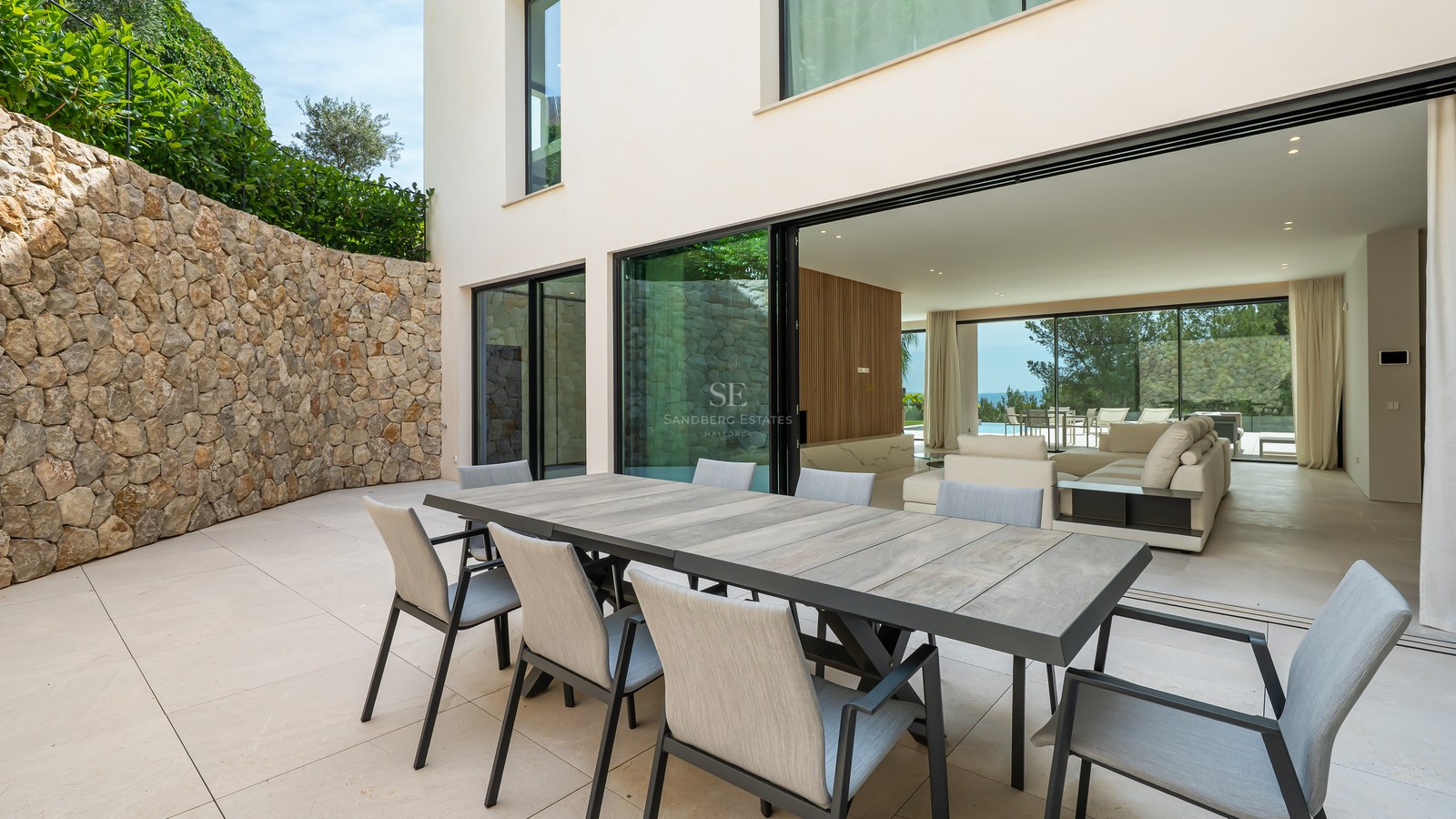Comedor exterior moderno en terraza de piedra con gran muro de piedra natural y puertas correderas de cristal.