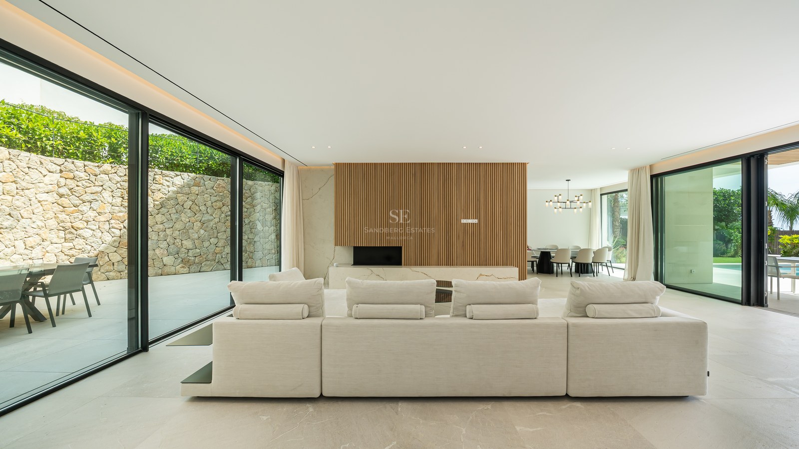 Amplio sofá modular blanco en un salón luminoso con grandes ventanales de cristal y paneles de madera.