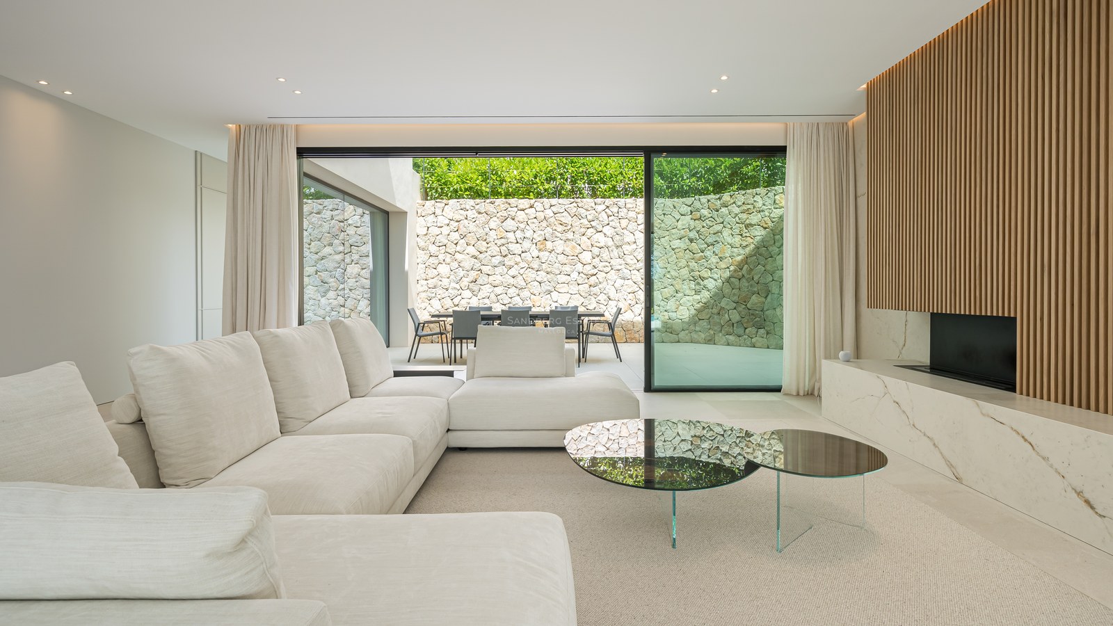 Elegante salón contemporáneo con sofá modular blanco, chimenea de mármol y ventanales que abren a un patio de piedra.