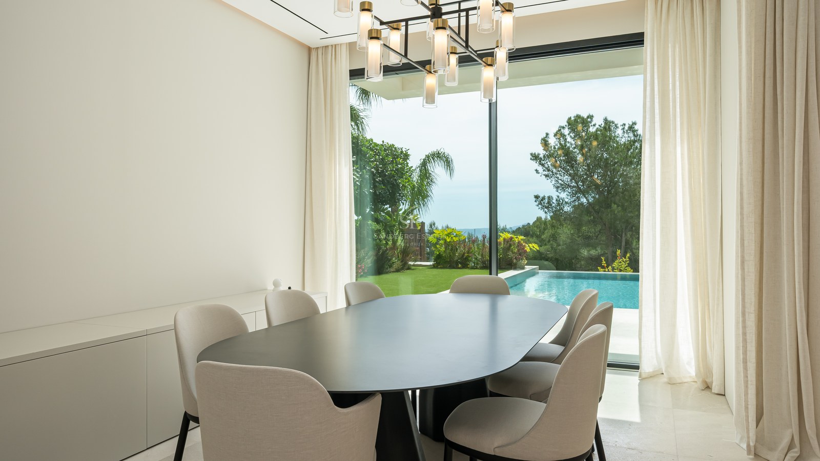 Comedor minimalista con mesa ovalada negra y ventanales de suelo a techo con vistas a una piscina y jardín.