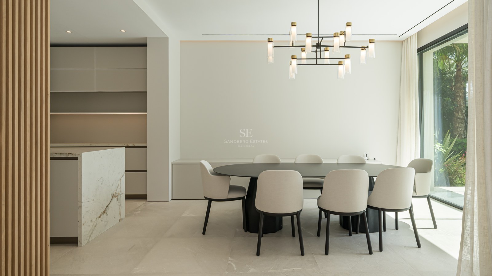 Comedor minimalista con mesa negra, sillas crema, isla de mármol y ventanal de suelo a techo hacia el jardín.