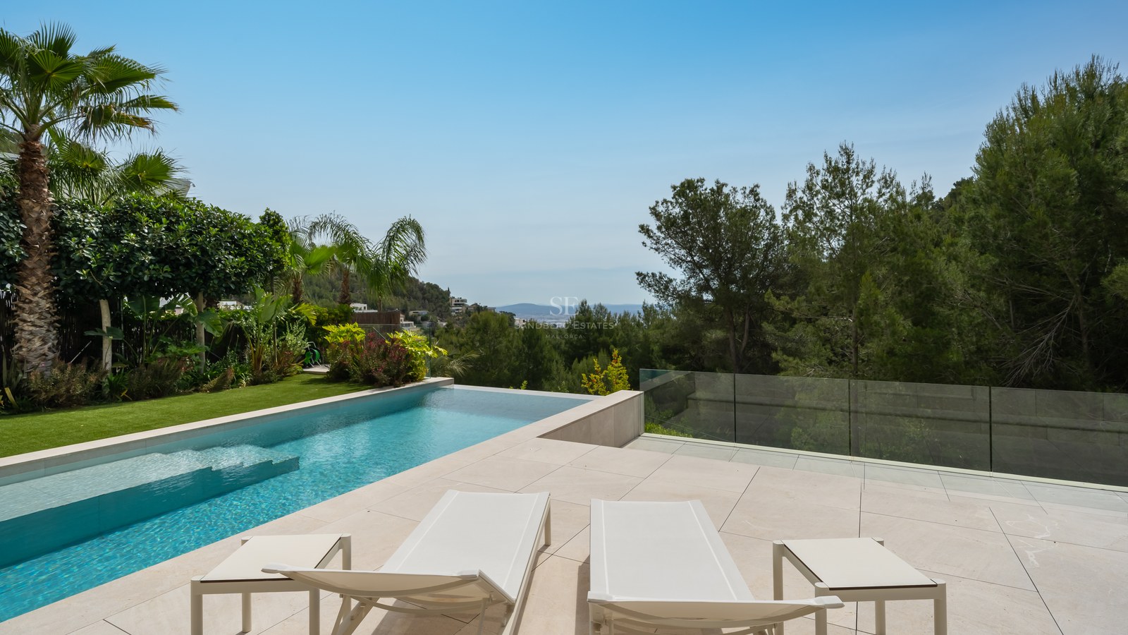 Piscina infinita moderna y terraza de piedra con hamacas blancas frente a un bosque de pinos y el Mediterráneo.
