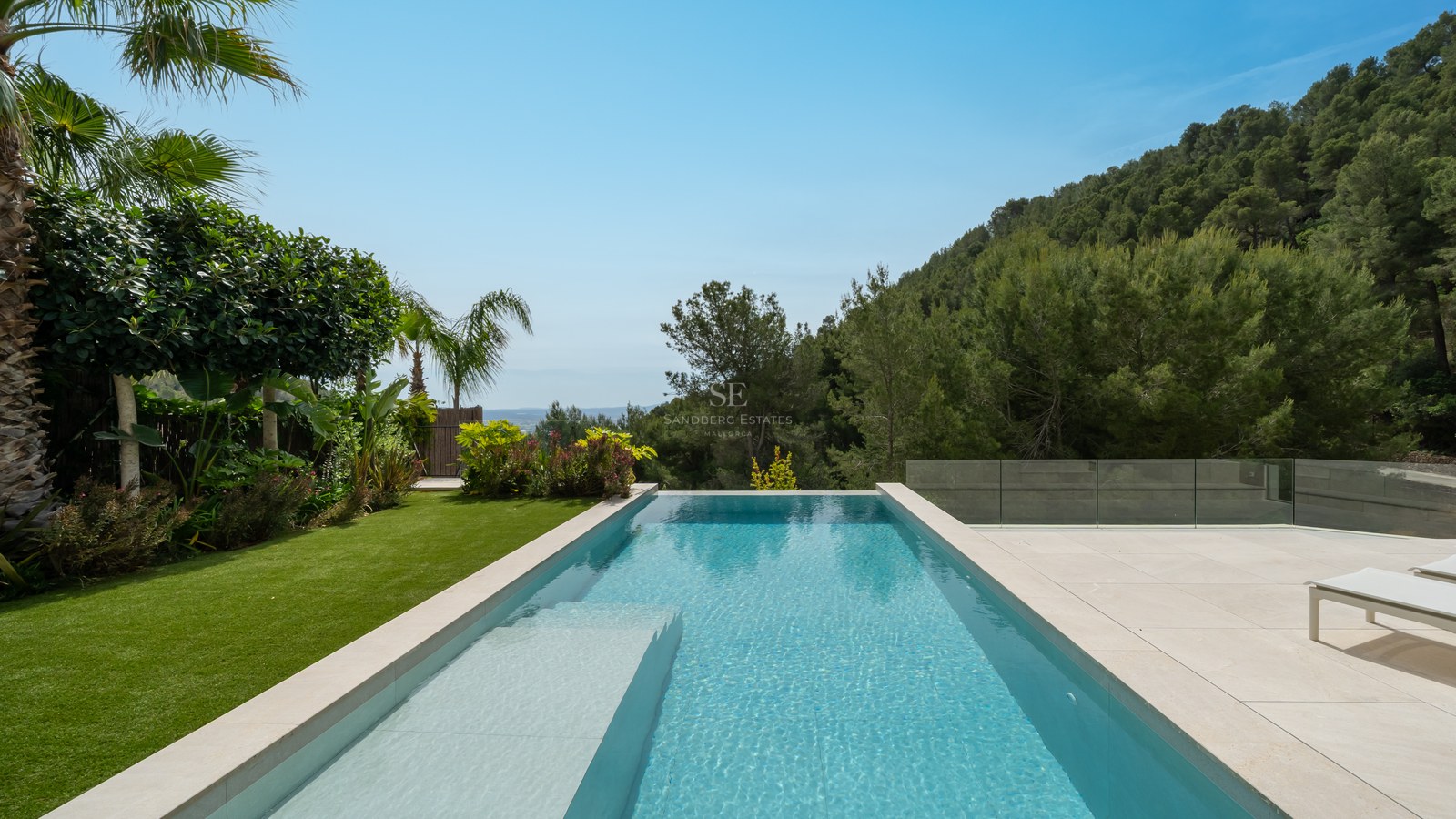 Piscina de borde infinito con suelo de piedra crema con vistas a un bosque de pinos y al mar de fondo.