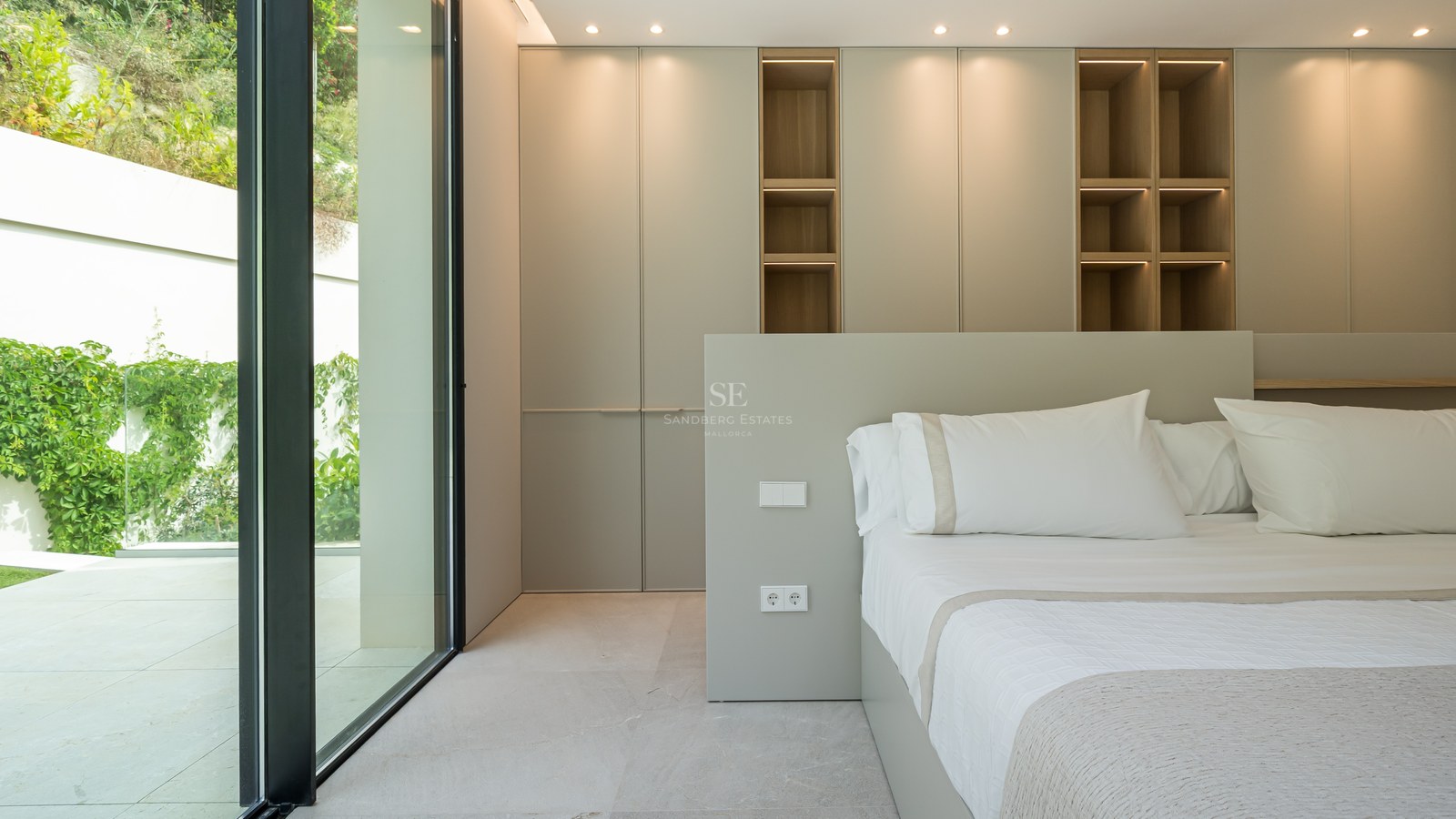 Dormitorio contemporáneo con ventanales de suelo a techo, armarios empotrados y estanterías de madera iluminadas.