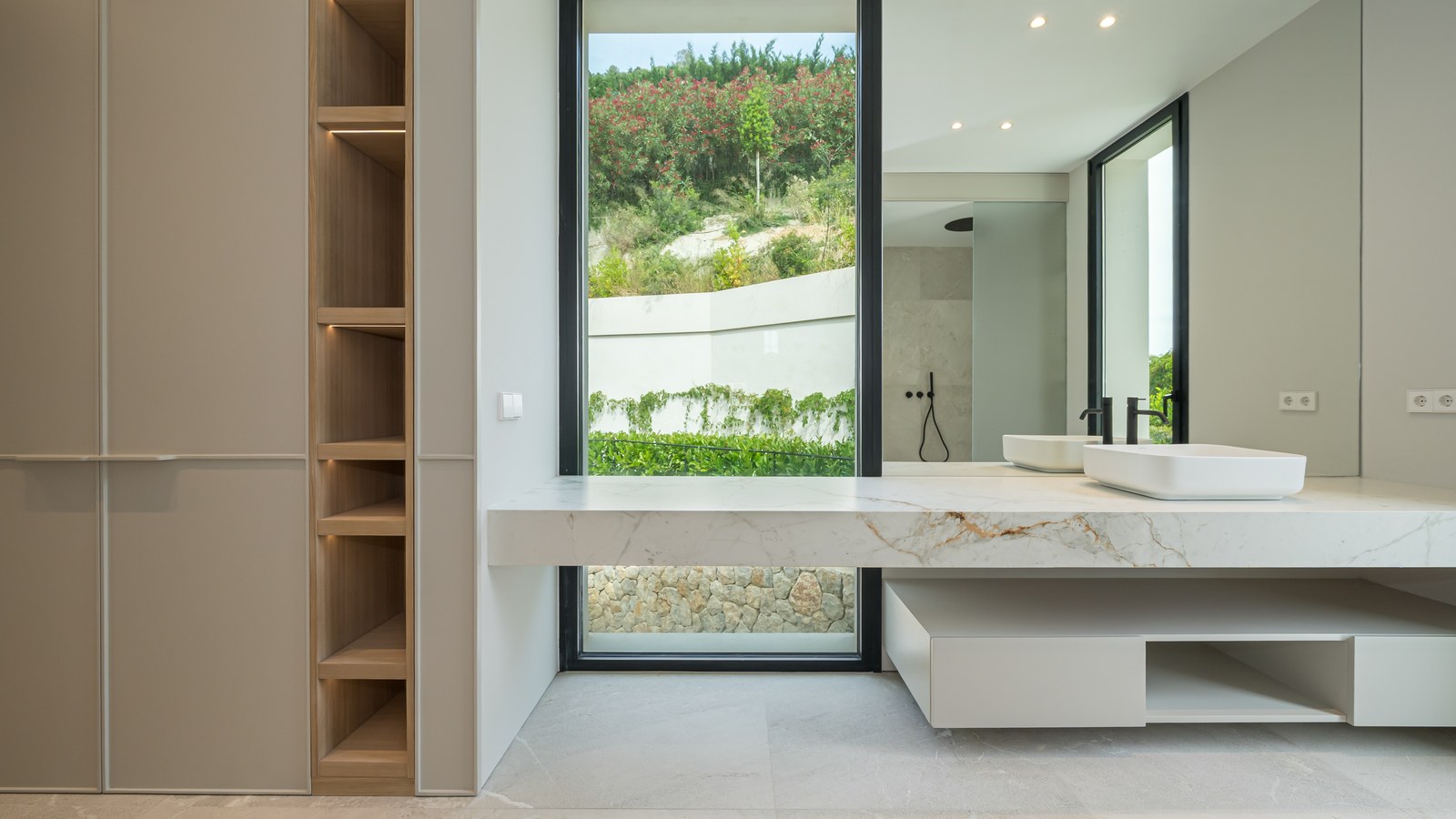 Baño moderno con encimera de mármol, lavabo sobre encimera, grifería negra y ventanal con vistas a la naturaleza.
