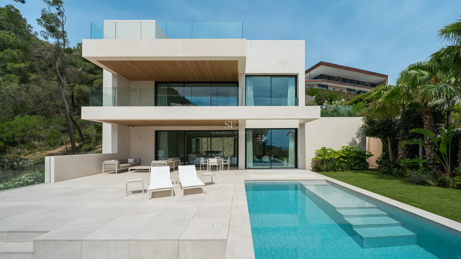 Vista trasera de una villa contemporánea de tres plantas con piscina turquesa, terraza de piedra y jardín.