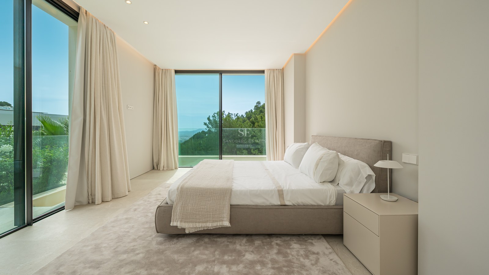 Dormitorio luminoso con cama doble, ventanales de suelo a techo y vistas a una colina arbolada bajo un cielo despejado.