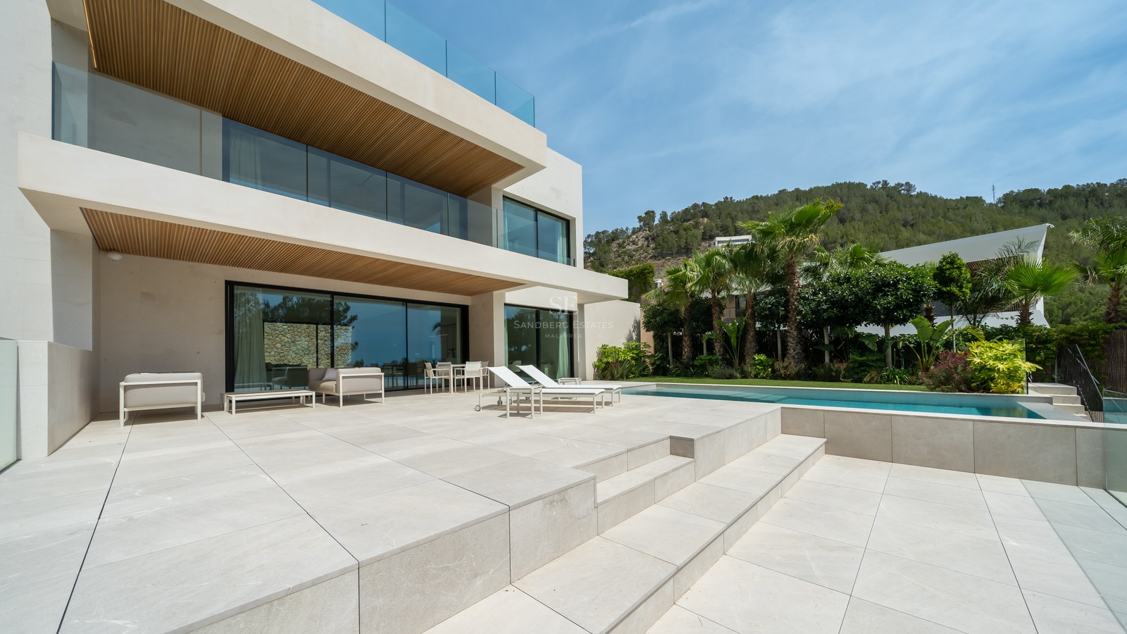 Villa moderna de piedra blanca con terraza amplia, piscina, barandillas de cristal y vistas a la montaña.