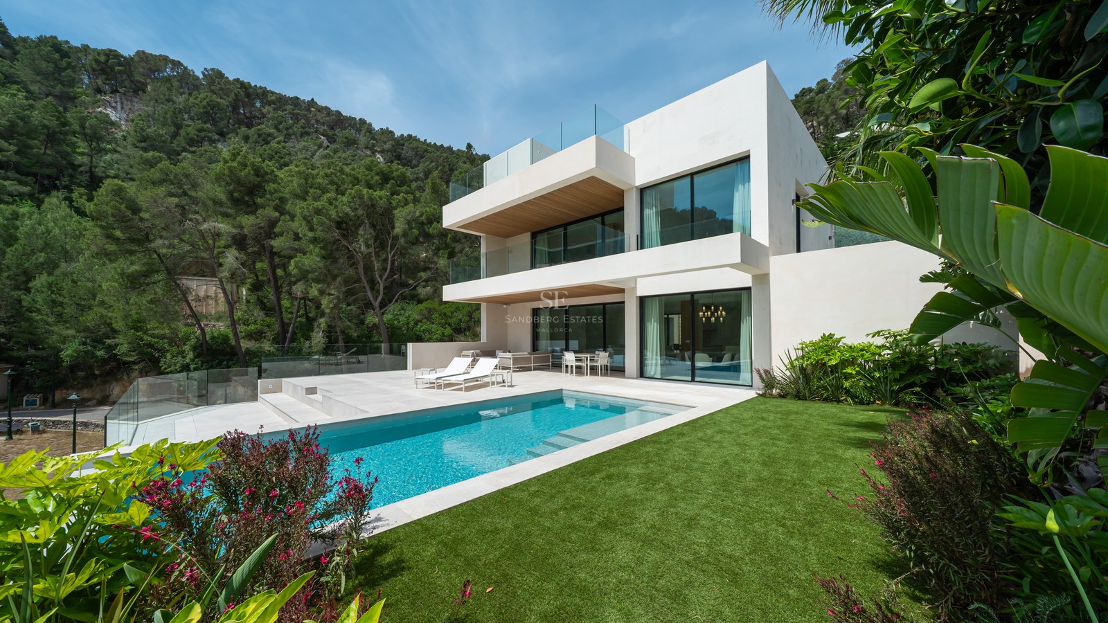 Villa blanca contemporánea con piscina rectangular, césped verde y balcones de cristal frente a una colina boscosa.