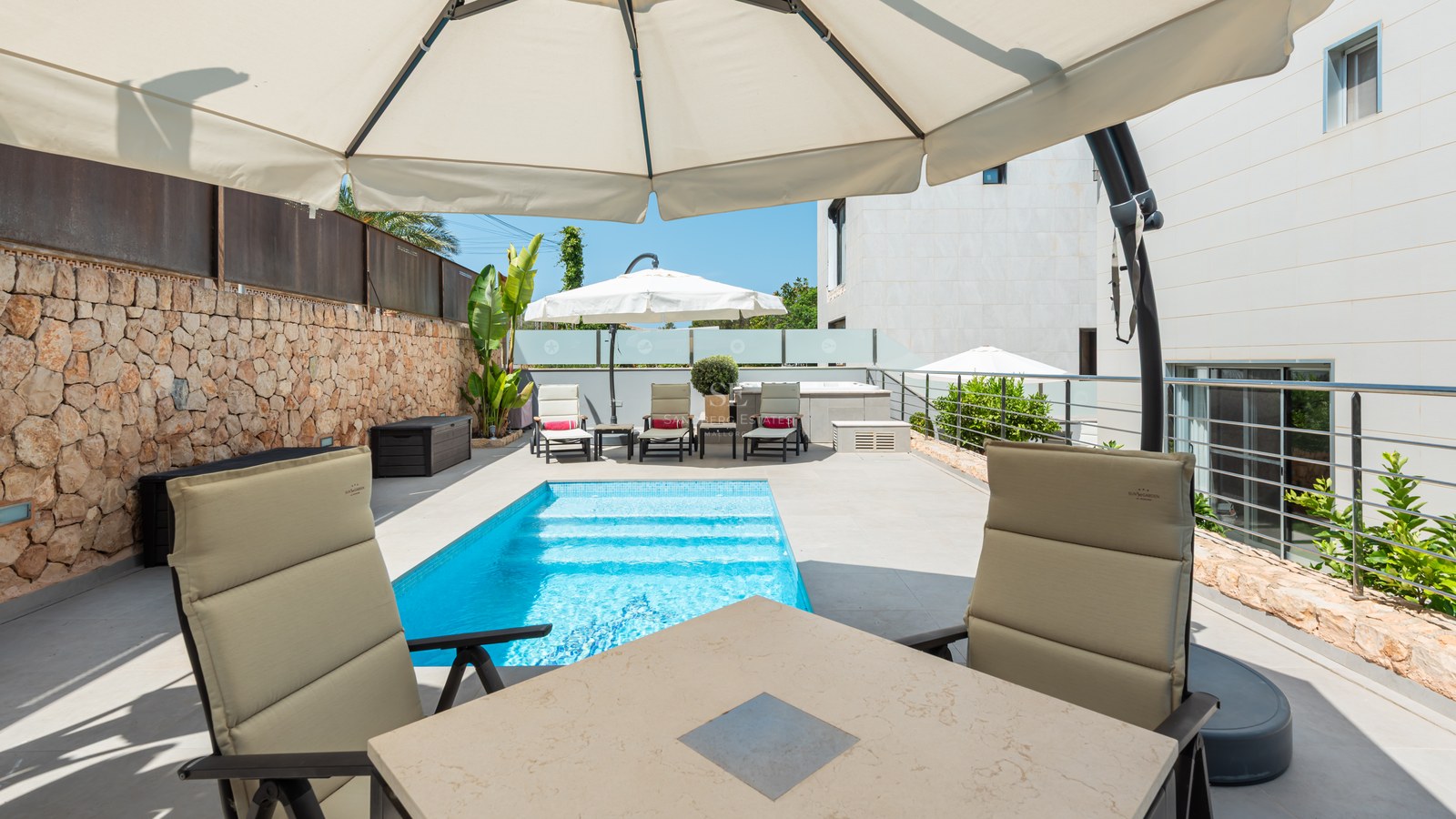 Piscina turquesa en terraza moderna con pared de piedra natural, tumbonas y sombrillas bajo el sol.