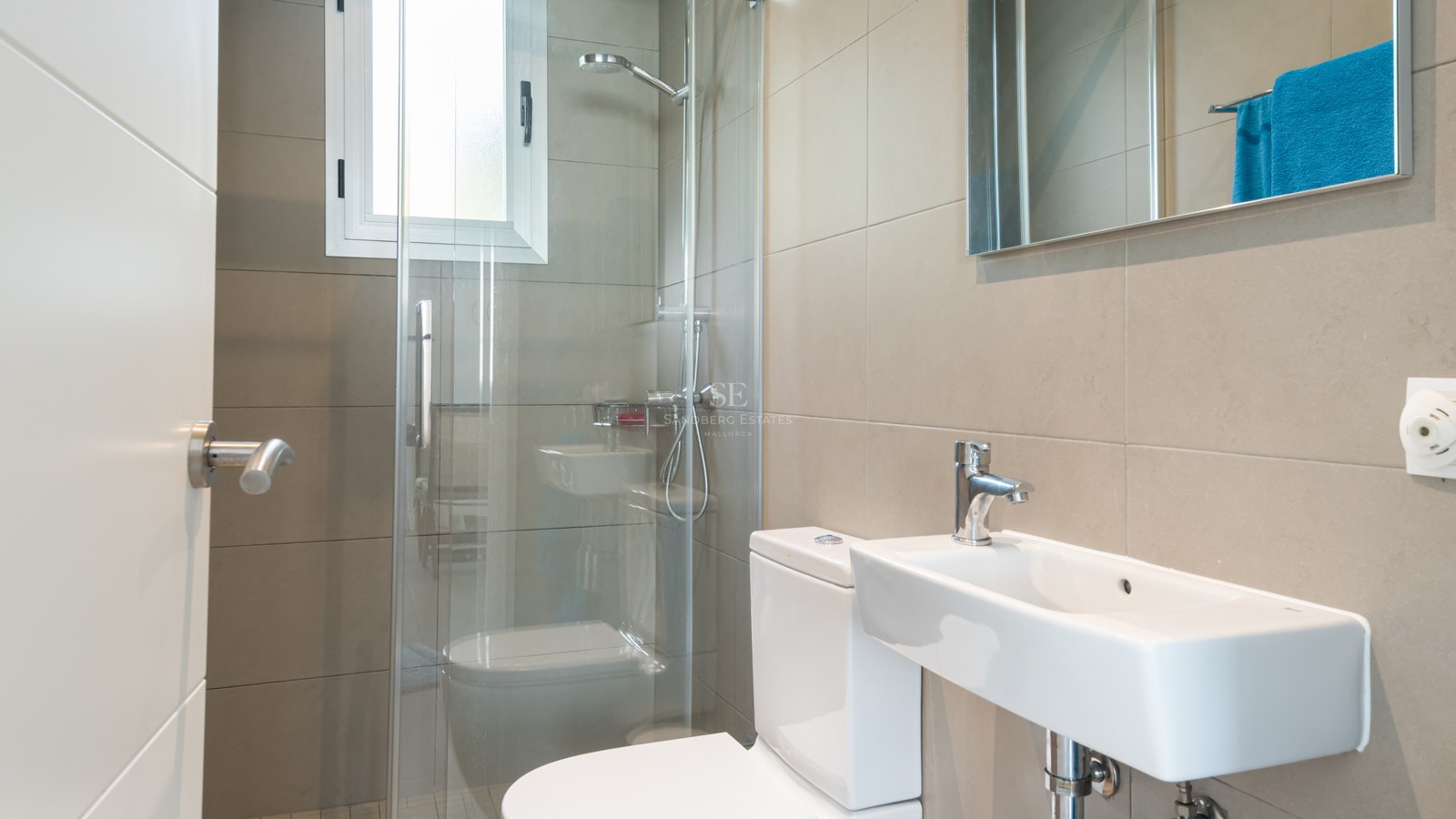 Baño contemporáneo con azulejos beige, ducha con mampara de cristal, lavabo blanco y luz natural.