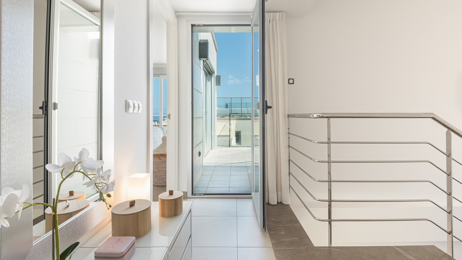 Ljus hall med vitt klinkergolv som leder till en öppen glasdörr och en solig terrass med metallräcke.