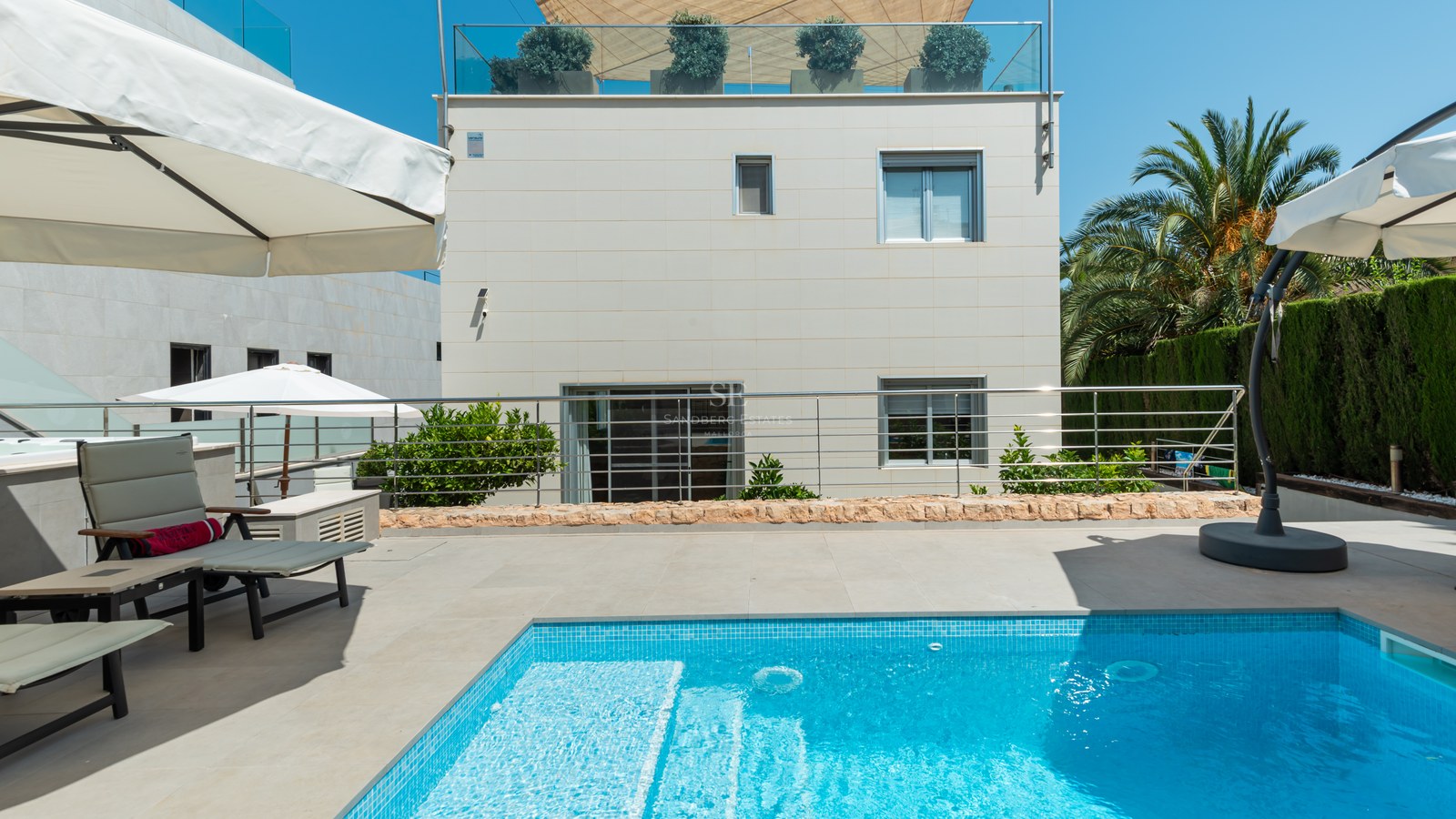 Privat utomhuspool med turkos mosaik, solstolar och vita parasoll framför en modern villafasad.