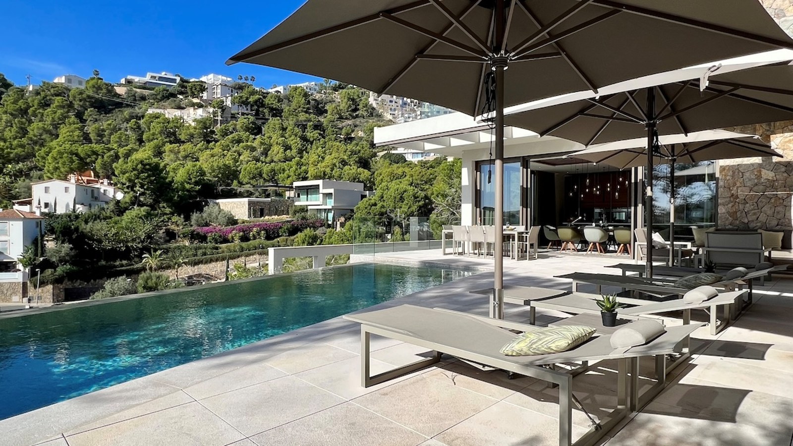 Vista exterior de villa moderna con piscina infinita, amplios ventanales y jardines exuberantes. Diseño minimalista y elegante.