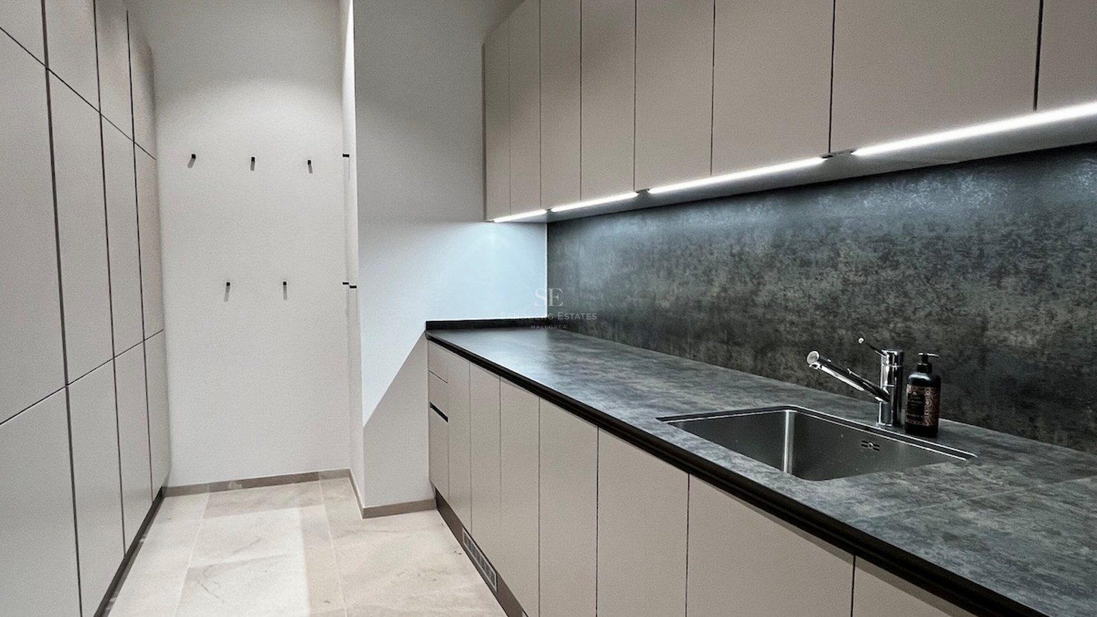 Lavandería moderna con encimera de piedra gris, muebles beige sin tiradores, iluminación LED y suelo de piedra.