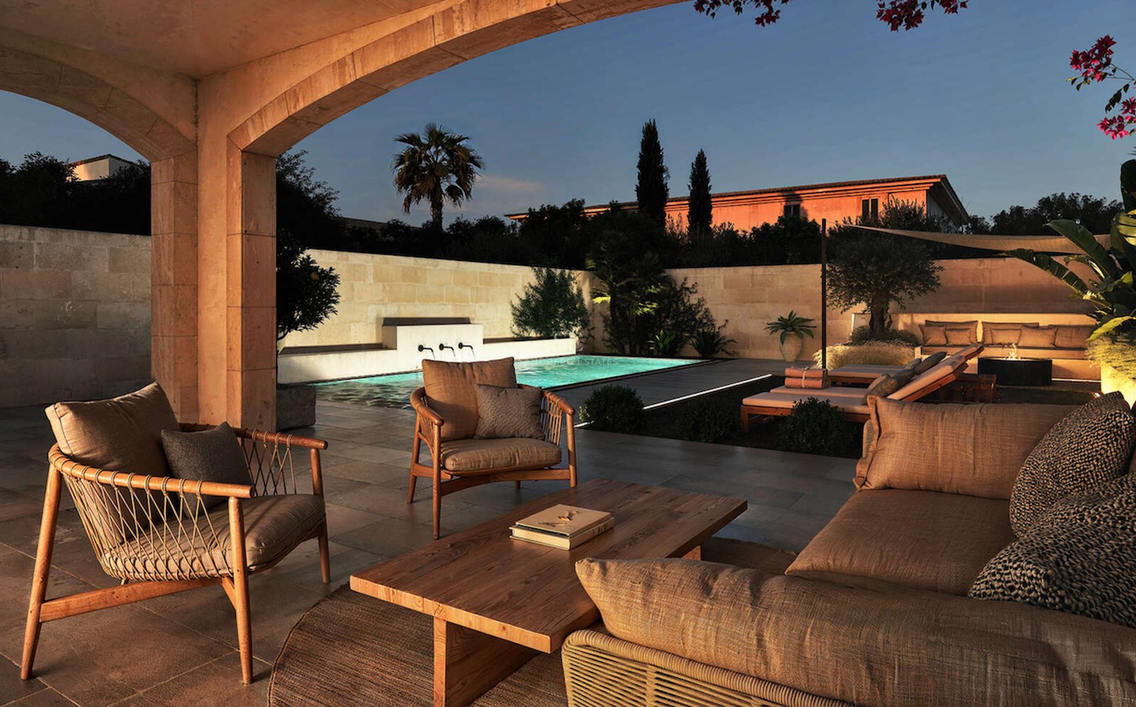 Salon extérieur avec mobilier en bois donnant sur une piscine illuminée et un jardin au coucher du soleil.