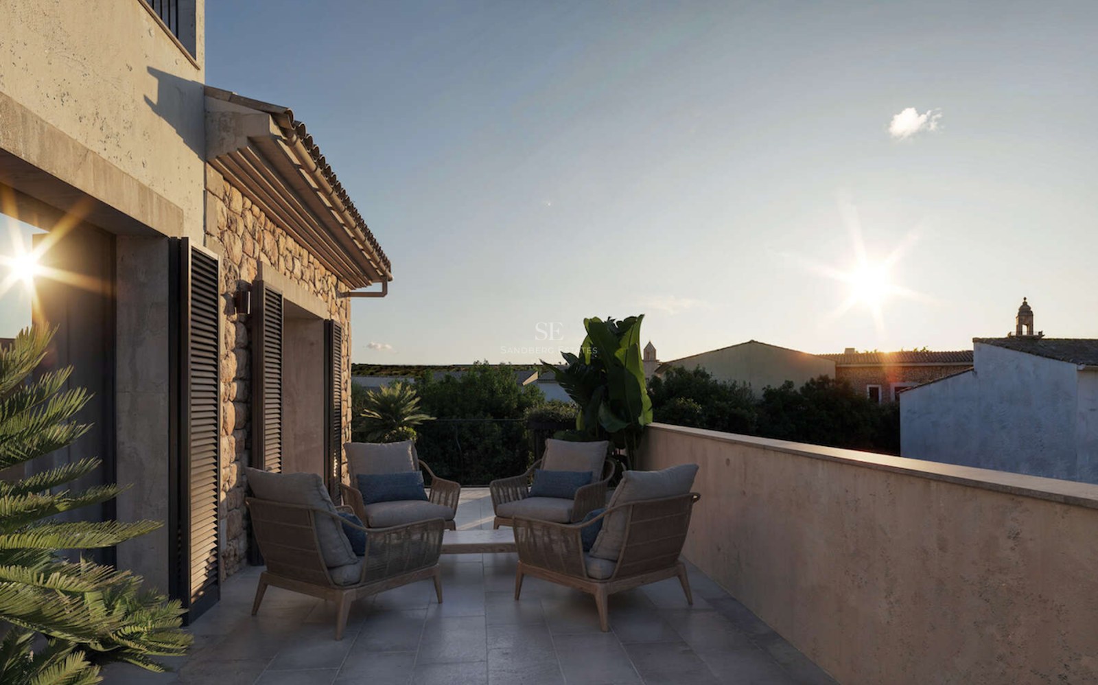 Terrasse avec fauteuils, murs en pierre naturelle et volets traditionnels surplombant un village au coucher du soleil.