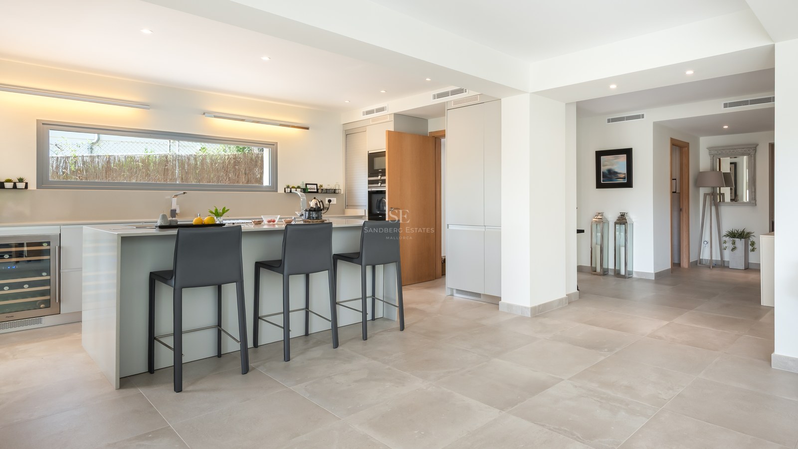 A contemporary kitchen featuring a large light grey island with three charcoal bar stools, a wine cooler, and tile flooring.
