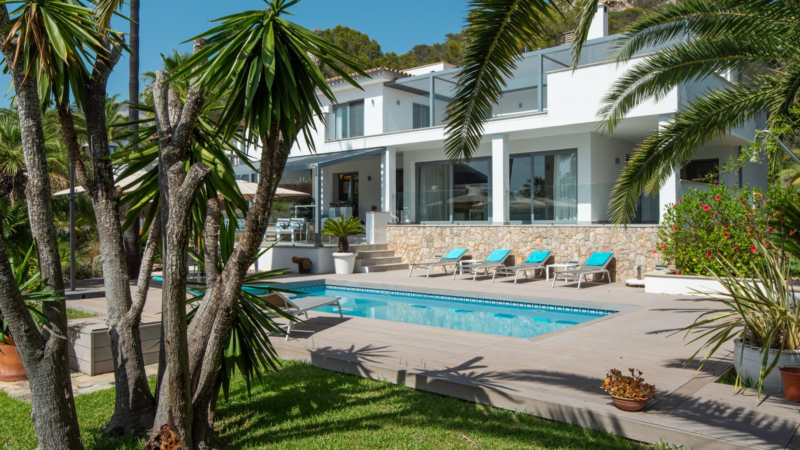 Modern white villa with a rectangular swimming pool, sun loungers, and lush palm trees under a clear blue sky.