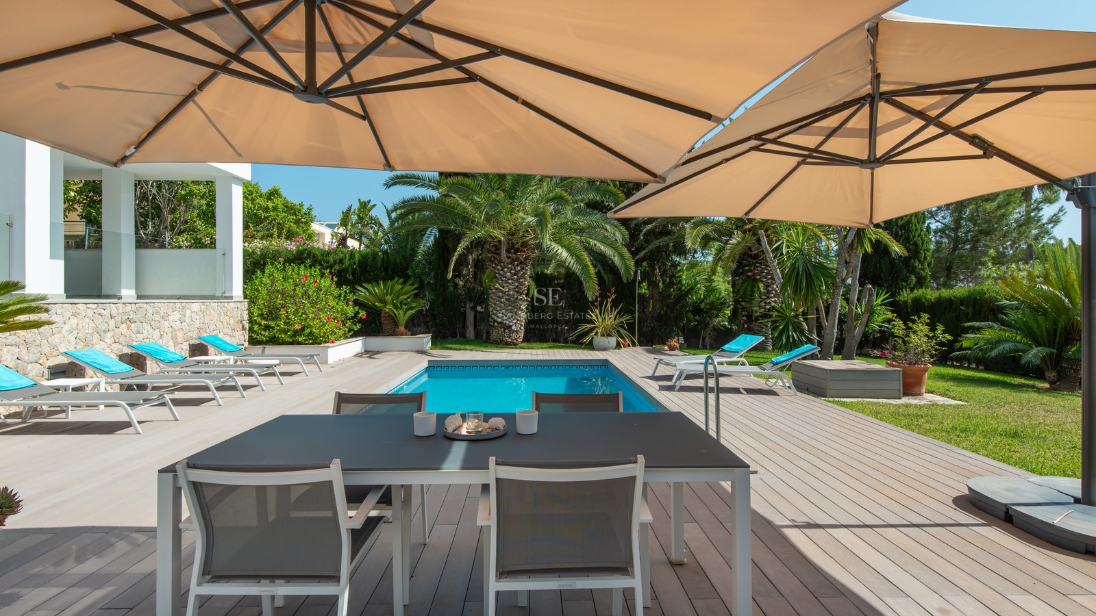 Wooden deck terrace featuring a dining table under large umbrellas next to a swimming pool and tropical palm trees.