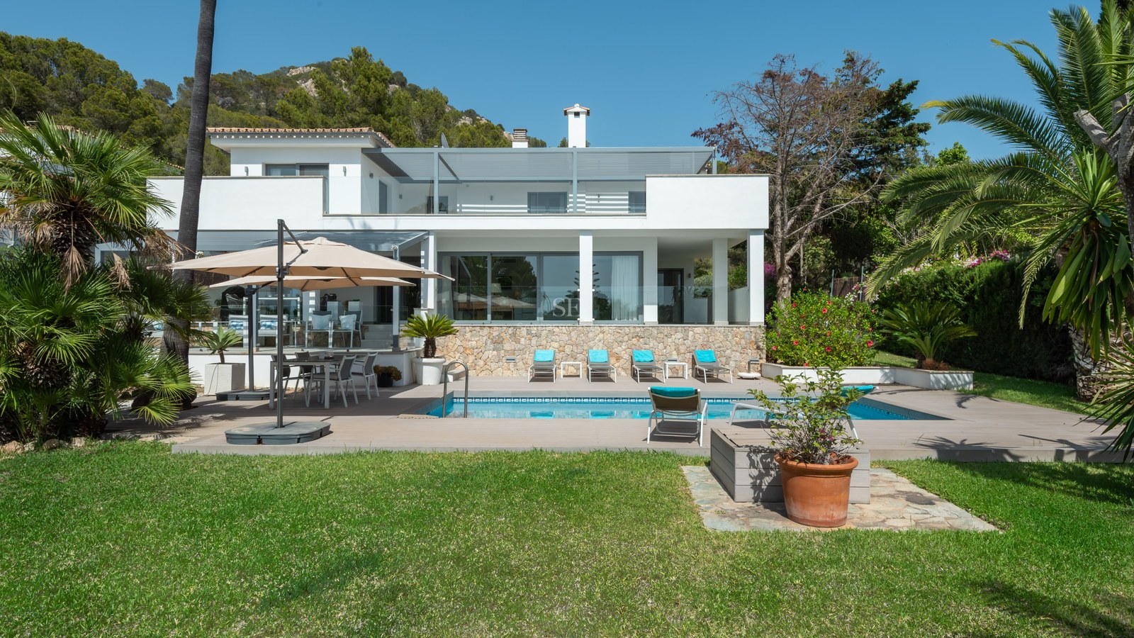 White contemporary villa featuring a swimming pool, sun loungers, and lush green lawn under a clear blue sky.