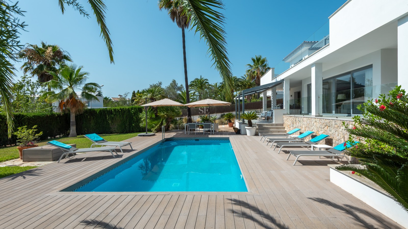 Rectangular swimming pool surrounded by wooden decking, white sun loungers, and palm trees under a clear blue sky.