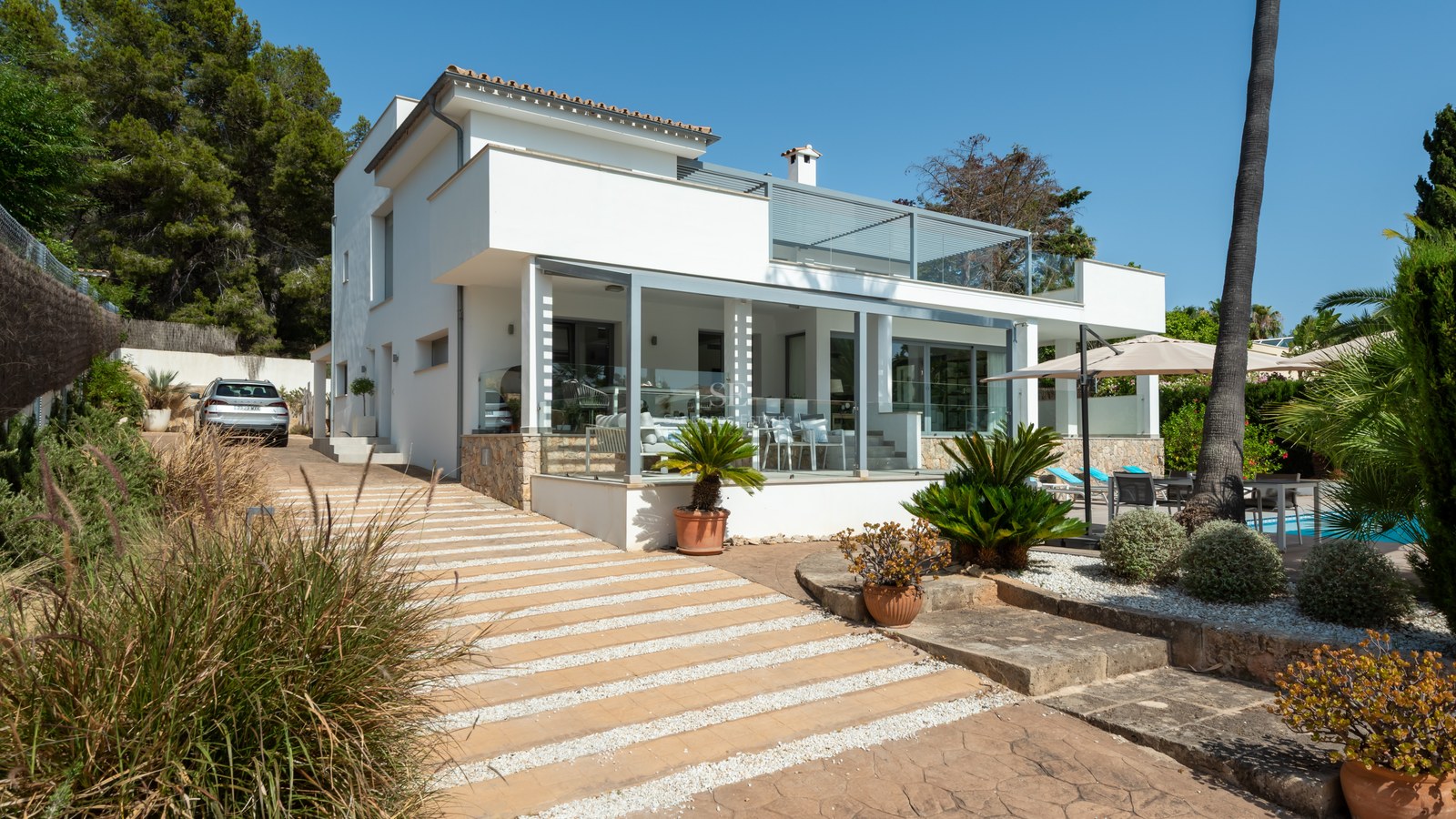 Two-story modern white villa with glass-fronted terraces, stone accents, and manicured landscaping under a clear blue sky.