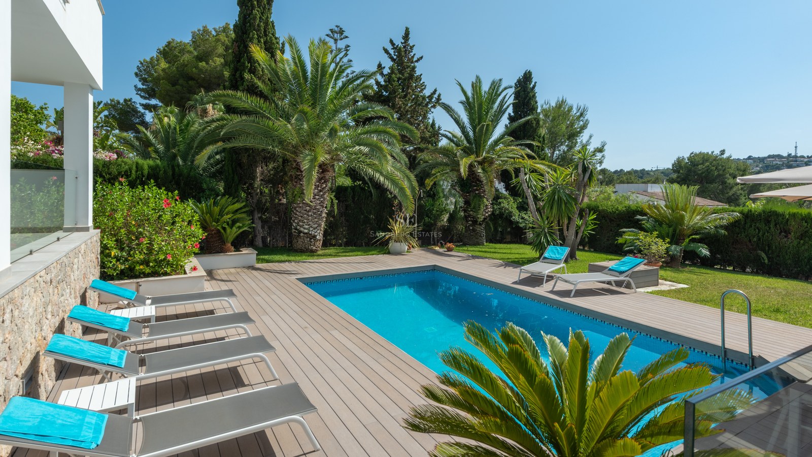 Rectangular blue pool surrounded by grey decking, sun loungers, and lush palm trees under a clear blue sky.