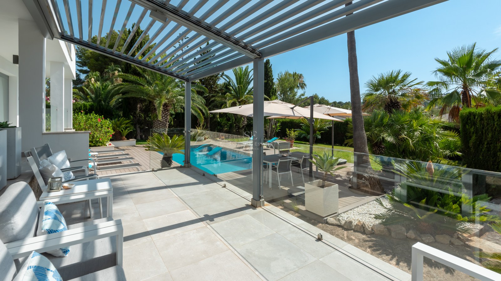 Modern covered terrace with lounge furniture overlooking a swimming pool and lush palm garden under a clear blue sky.