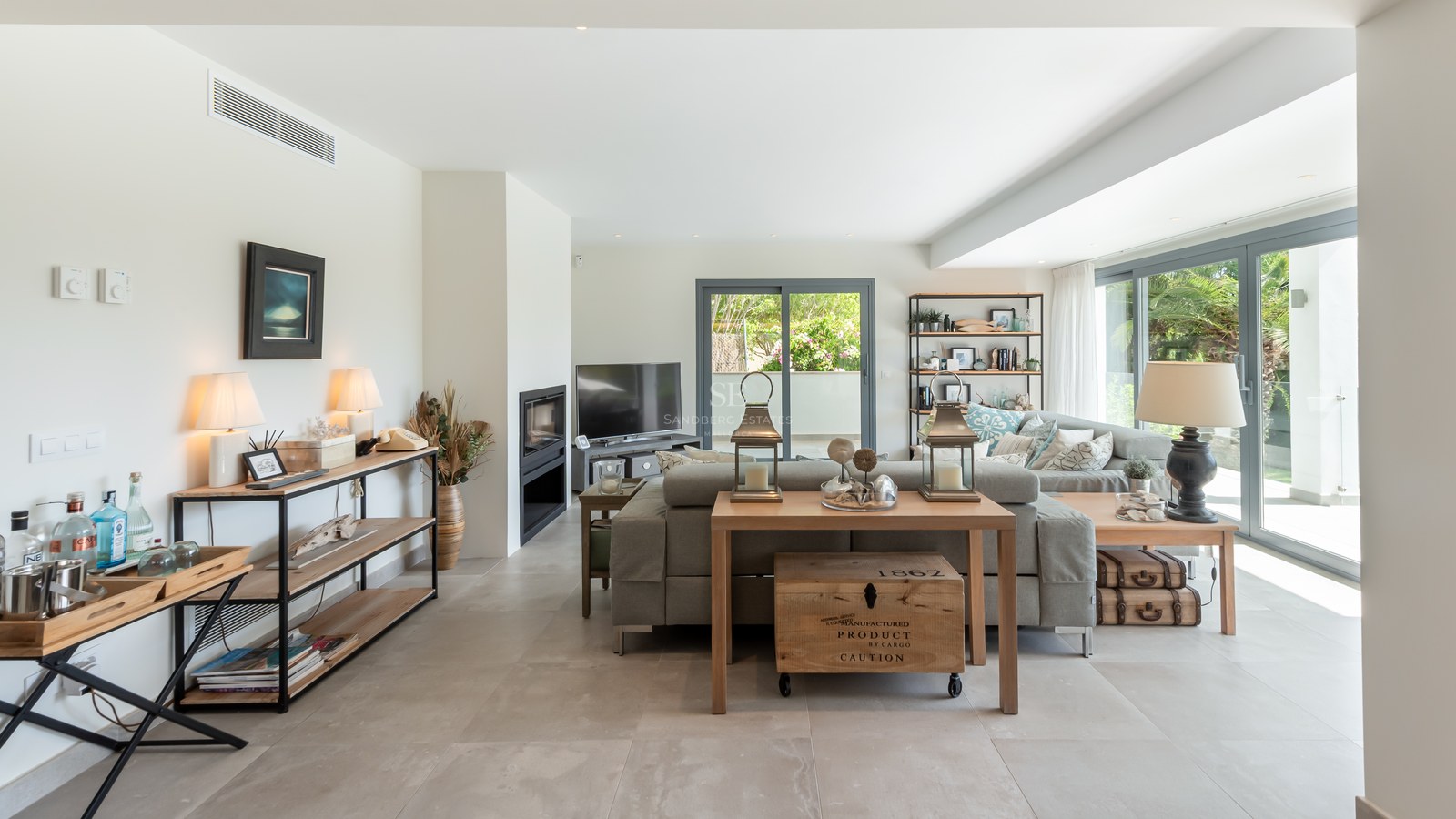 Spacious modern living room with grey sofa, large glass doors to garden, tile floors, and a built-in fireplace.