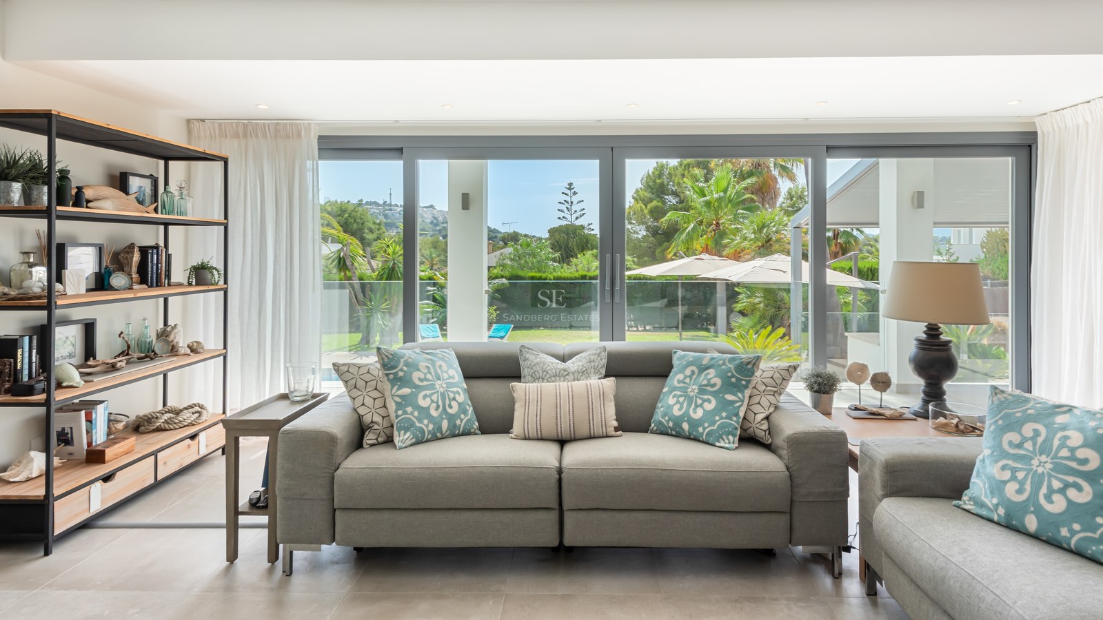 Modern grey sofa and industrial shelving in a bright living room with floor-to-ceiling glass doors opening to a garden.