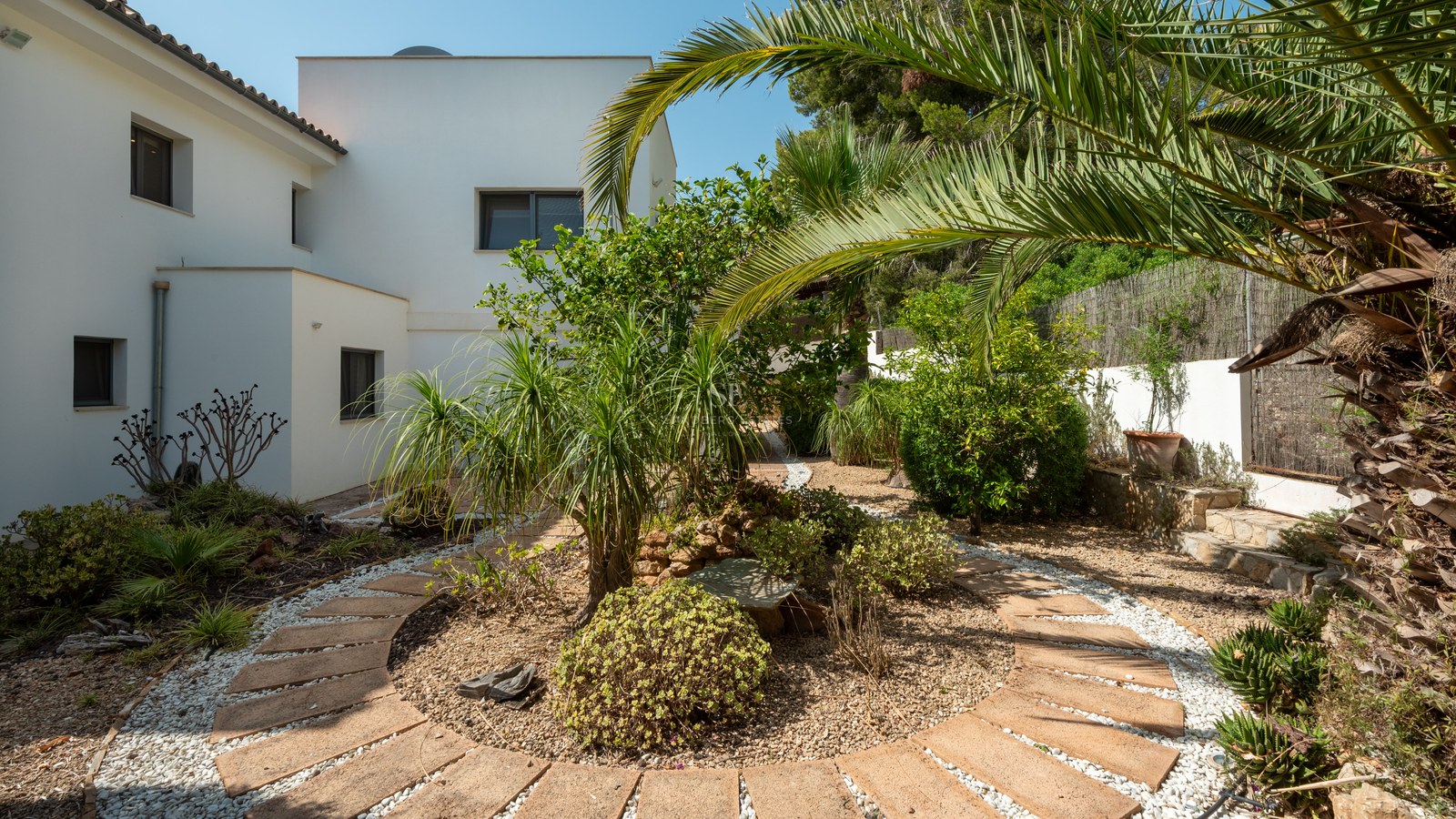 Circular stone pathway in a Mediterranean garden with palm trees and a white modern villa in the background.