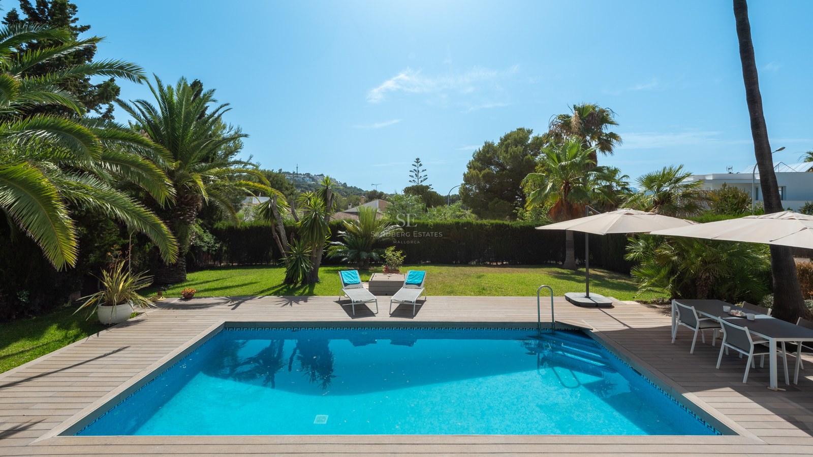 A rectangular turquoise pool surrounded by a wooden deck, sun loungers, and lush palm trees under a bright blue sky.