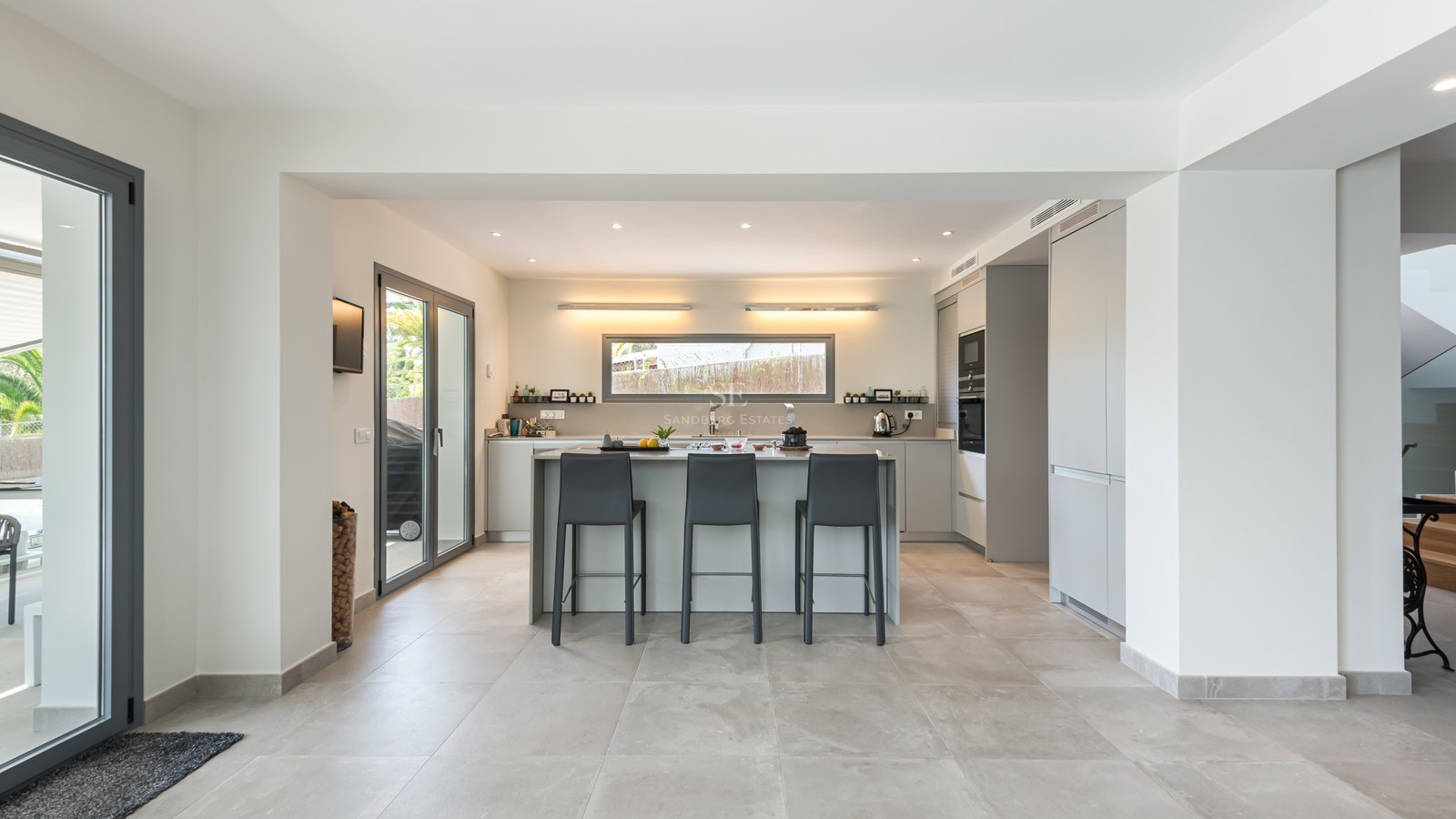 A minimalist grey and white kitchen featuring a large central island with charcoal bar stools and porcelain tile floors.