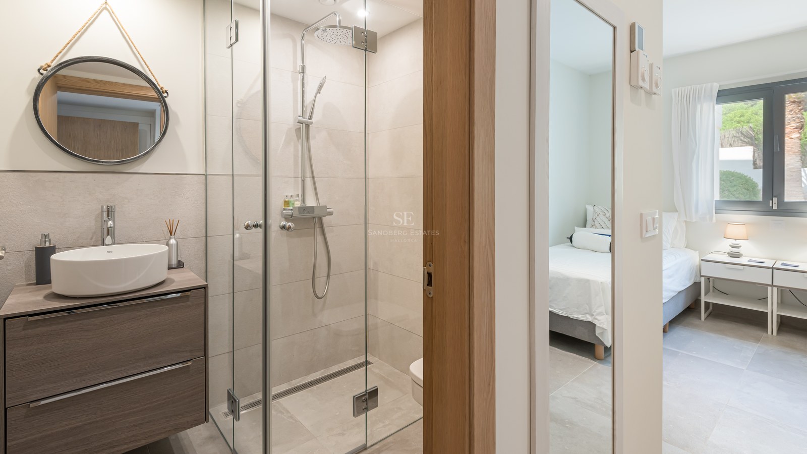 Modern bathroom featuring a glass walk-in shower, ceramic vessel sink, and round mirror, looking into a bedroom.