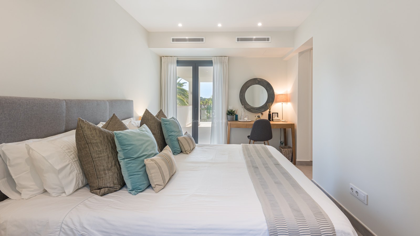 Bright bedroom featuring a large bed with teal accents, a wooden vanity desk, and floor-to-ceiling glass doors leading to a terrace.