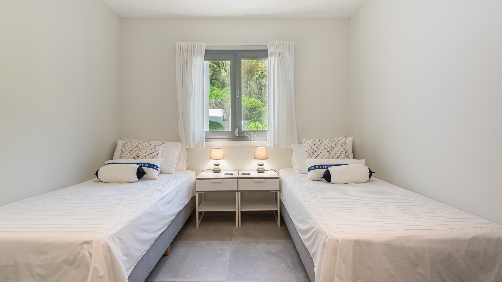 Bright guest bedroom featuring two single beds, white linens, and a window overlooking lush green gardens.