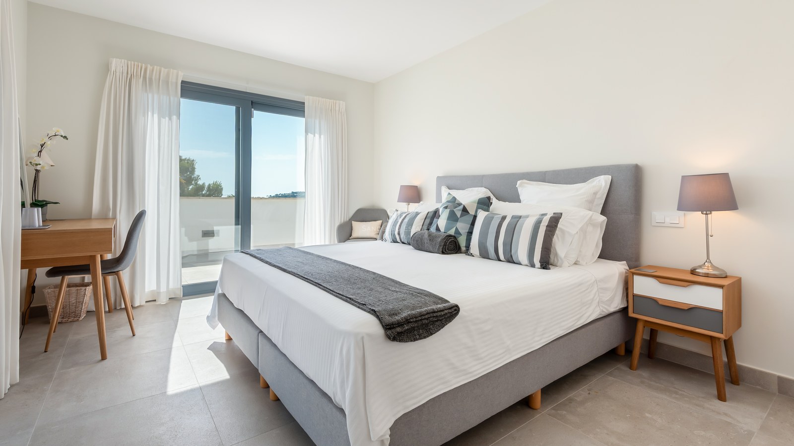 Bright bedroom with gray upholstered bed, wooden desk, and sliding glass doors to a sunny terrace.