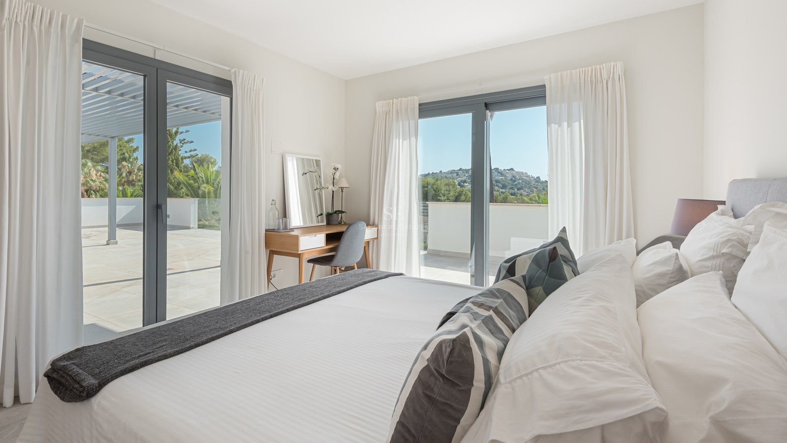 Bright contemporary bedroom with large glass doors opening onto a spacious tiled terrace with hillside views.