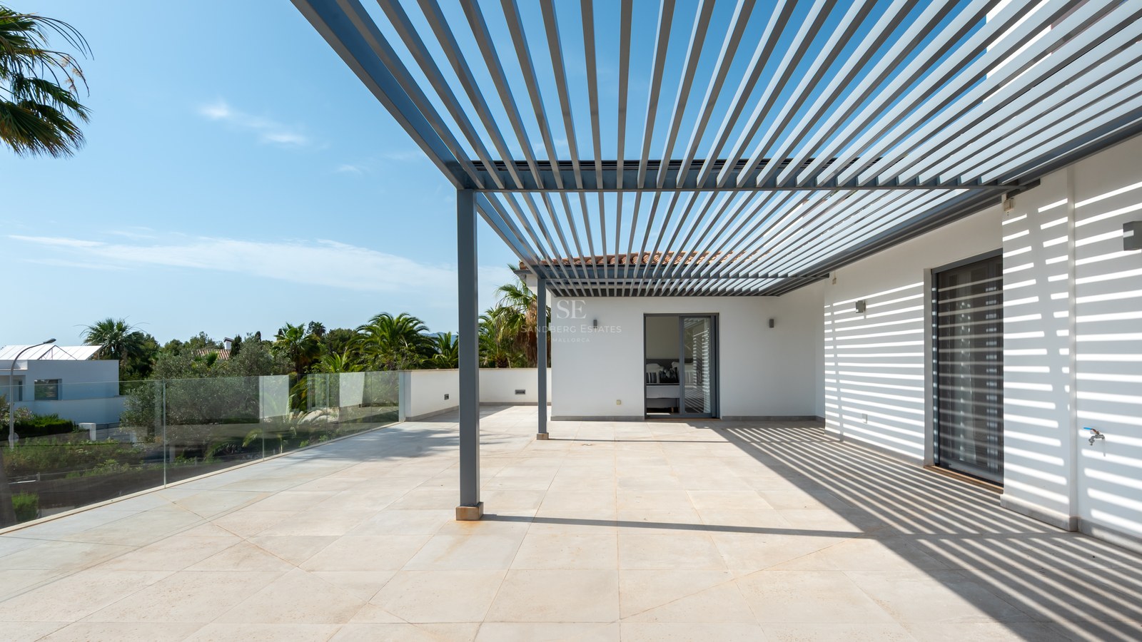 Large sun-drenched terrace with a gray metal pergola, glass railings, and views of palm trees under a blue sky.