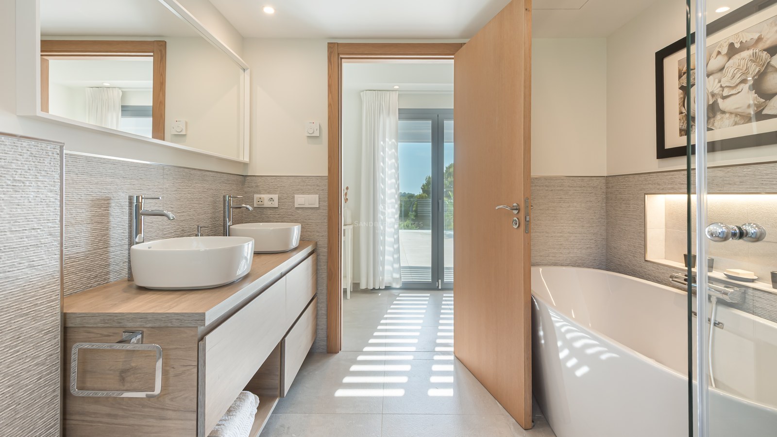 Modern bathroom featuring double vessel sinks on a wooden vanity, a large bathtub, and textured beige wall tiles.