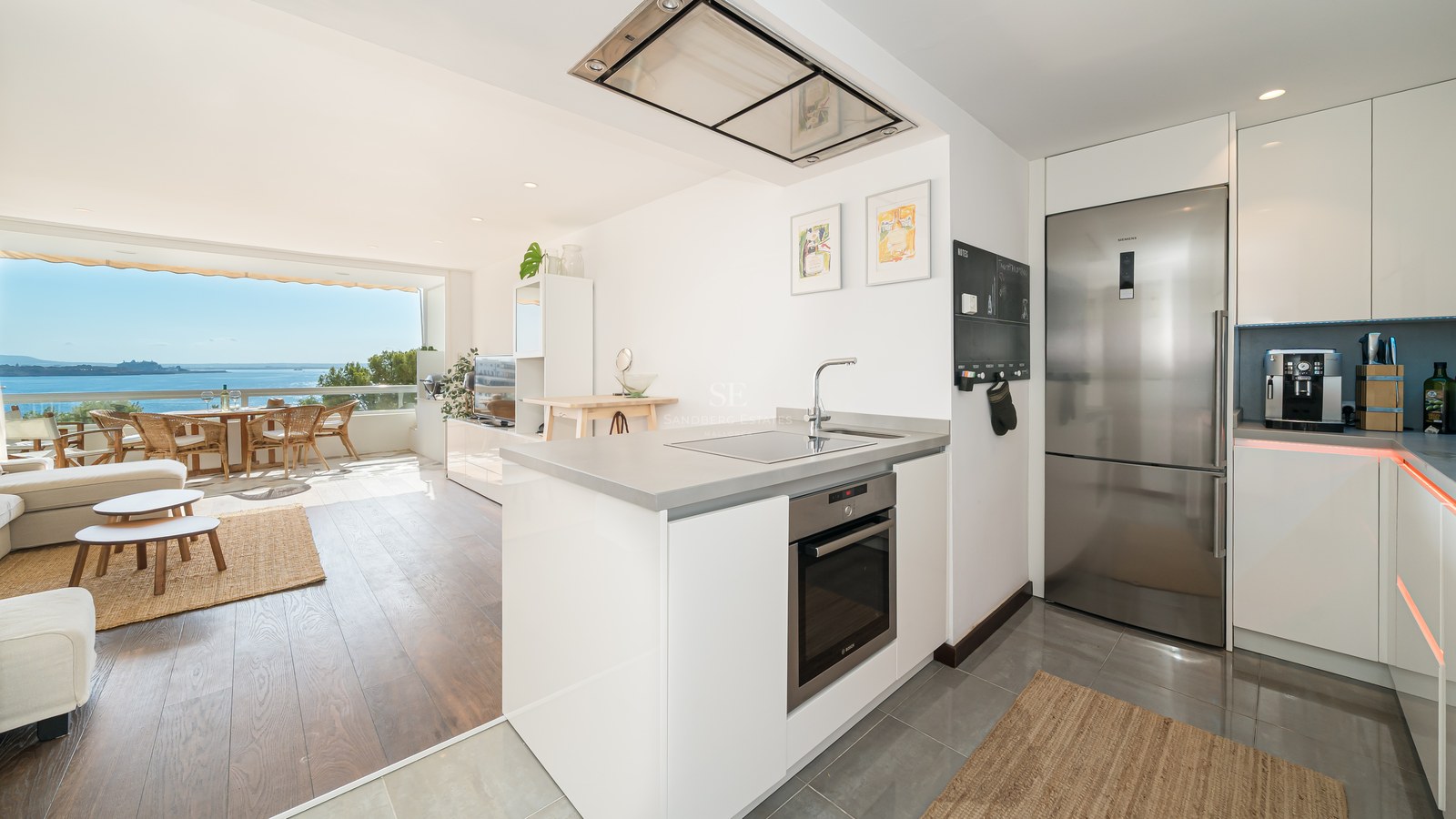 Cocina blanca moderna con electrodomésticos de acero inoxidable, salón y terraza con vistas al mar Mediterráneo.