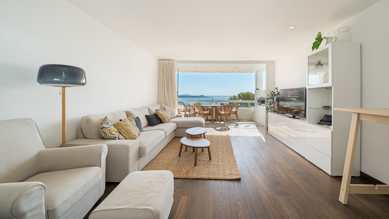 Salón luminoso con sofá grande, suelos de madera y puertas de cristal hacia terraza con vistas al mar.