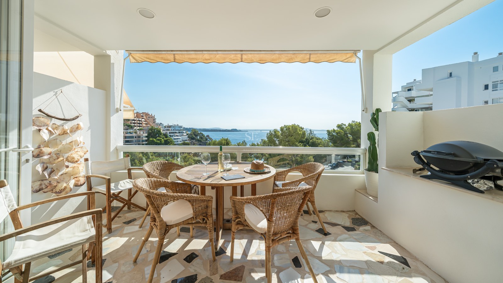 Terraza cubierta con mesa de comedor de madera, sillas de mimbre y barandilla de cristal con vistas al mar.
