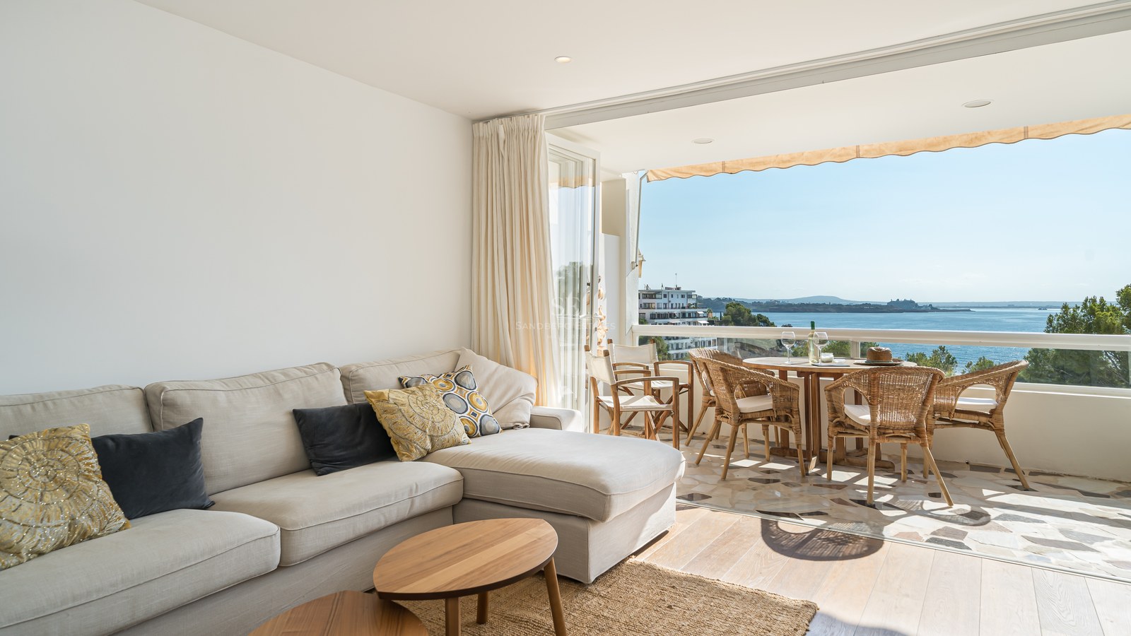 Salón interior con sofá beige que da a una terraza soleada con comedor y vistas al mar Mediterráneo.