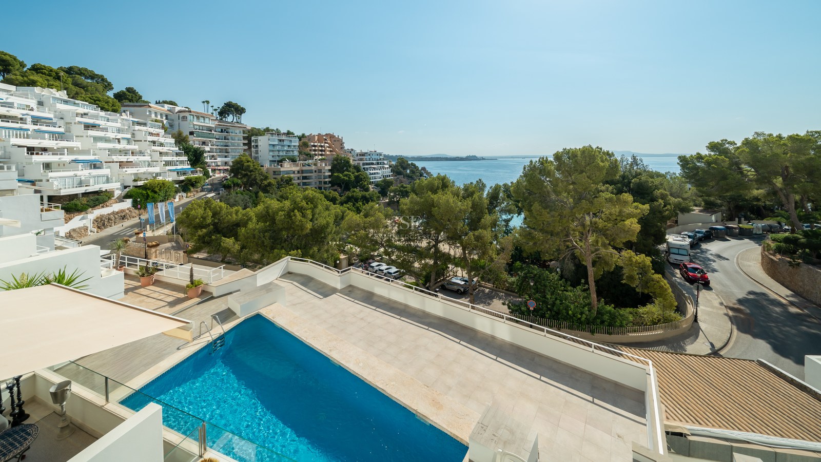 Terraza elevada con piscina rectangular azul y barandillas de cristal con vistas a una bahía mediterránea y pinos.