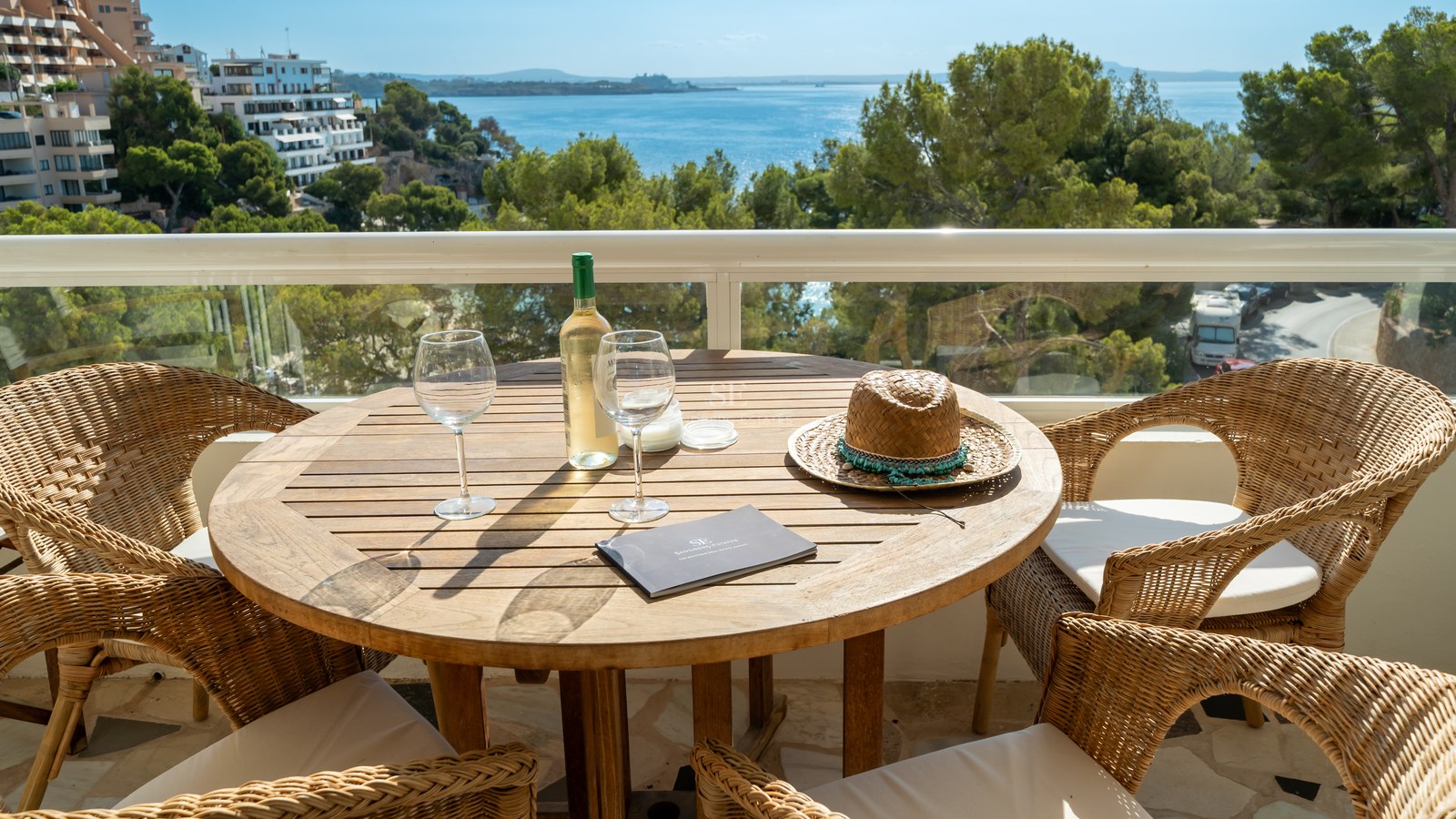 Terraza elevada con mesa de madera, sillas de mimbre y vistas al mar azul entre pinos mediterráneos.