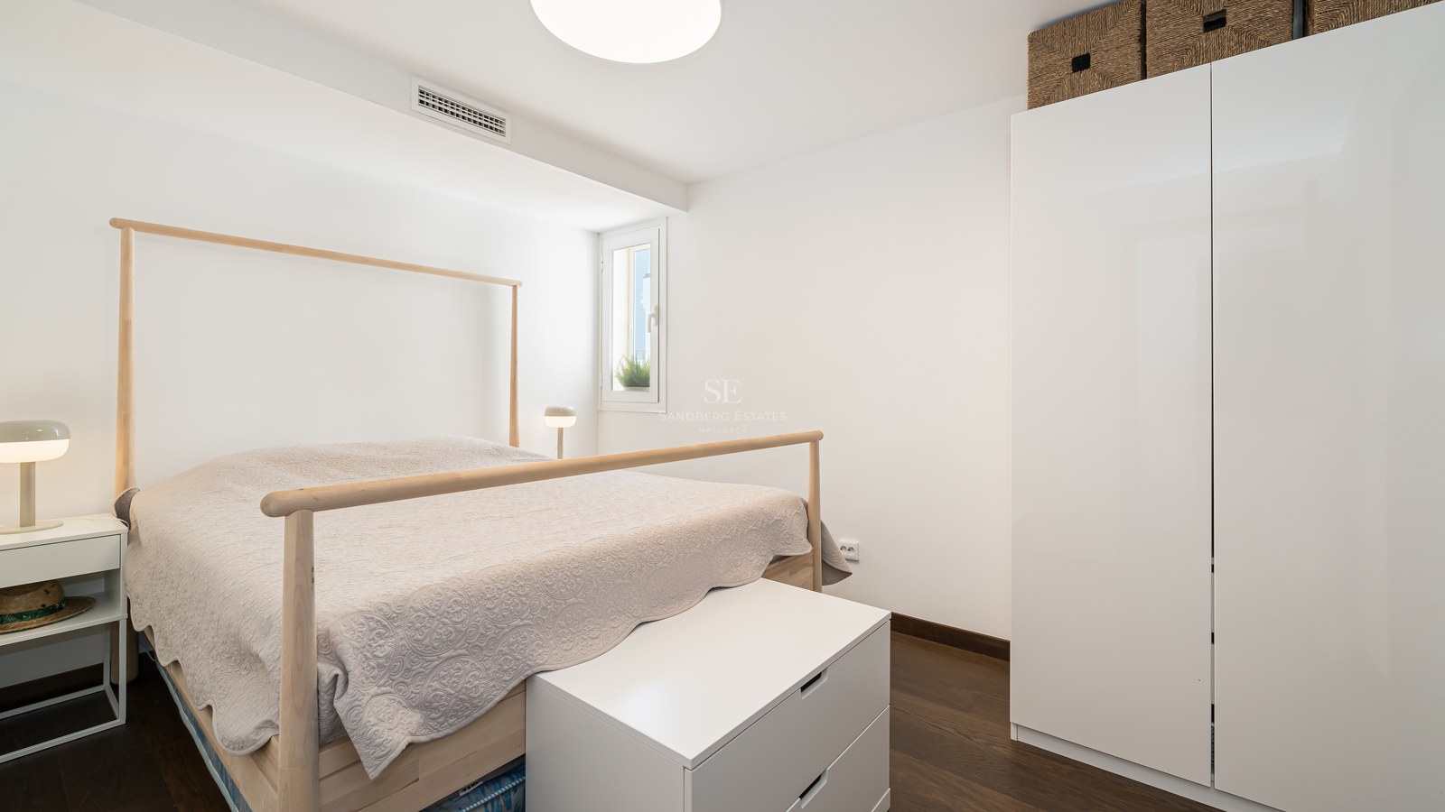 Habitación luminosa con estructura de cama de madera clara, armarios blancos y suelo de madera oscura.