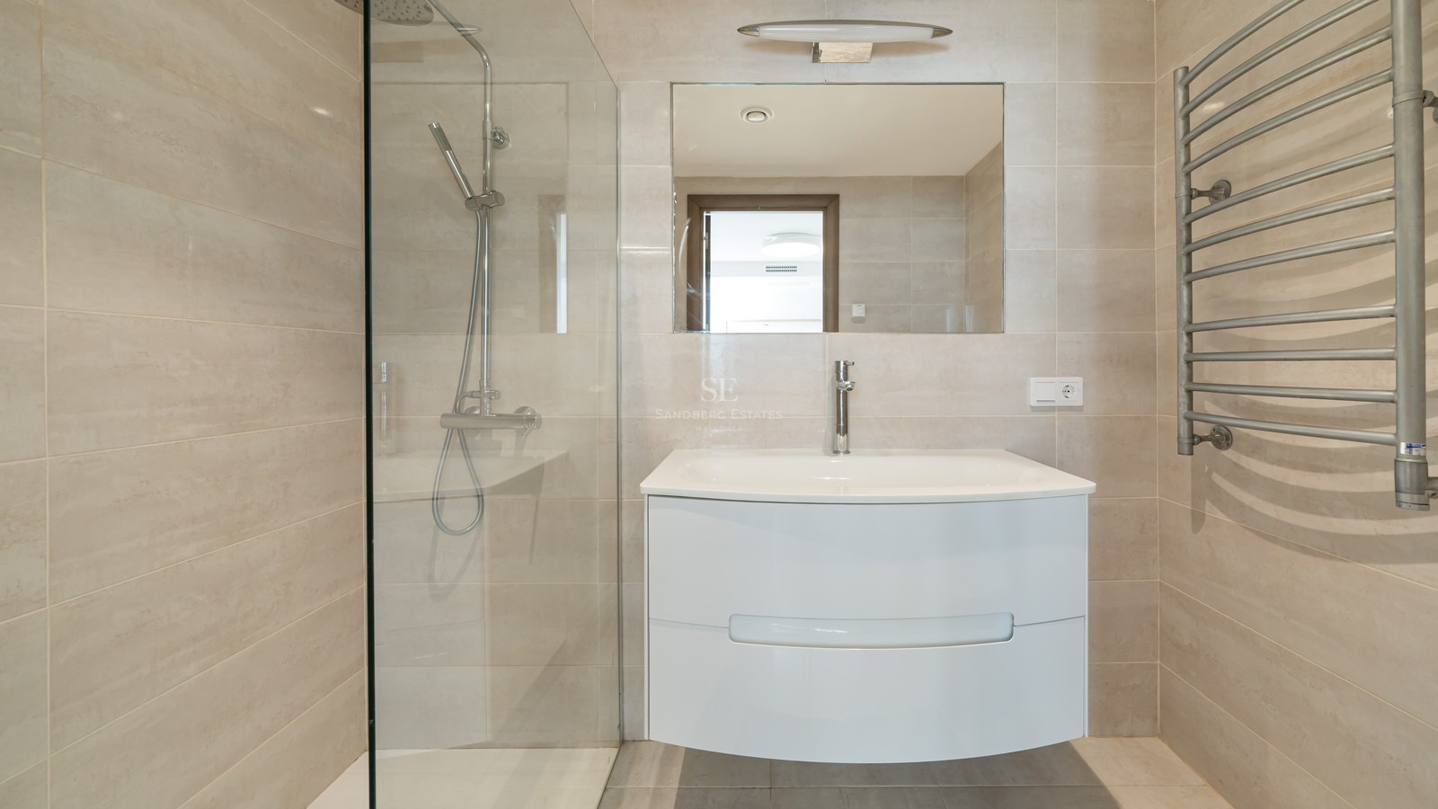 Baño moderno con azulejos beige, ducha con mampara de cristal, lavabo suspendido blanco y toallero calefactable.